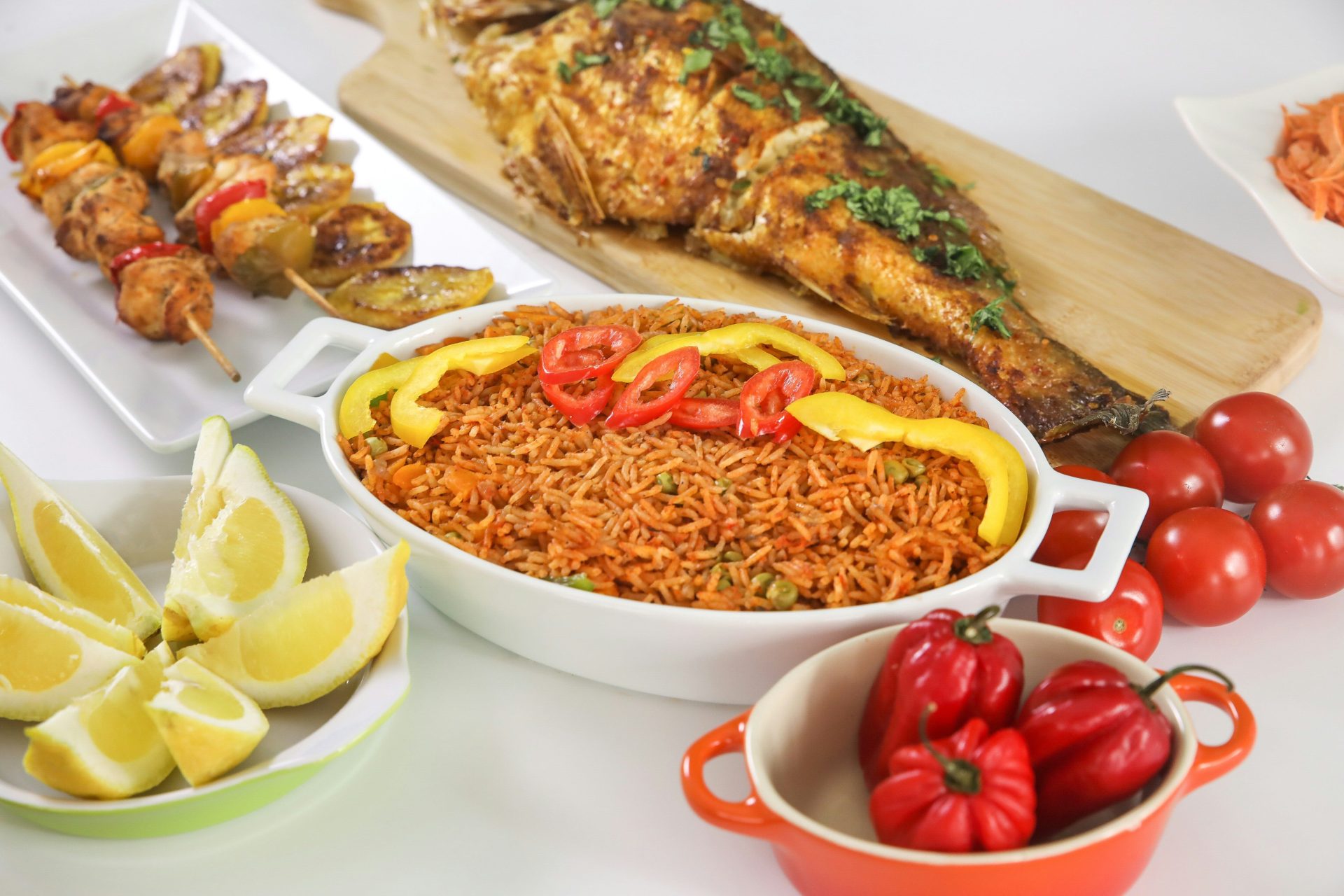 Jollof rice in a white dish with grilled fish, skewers, and fresh vegetables